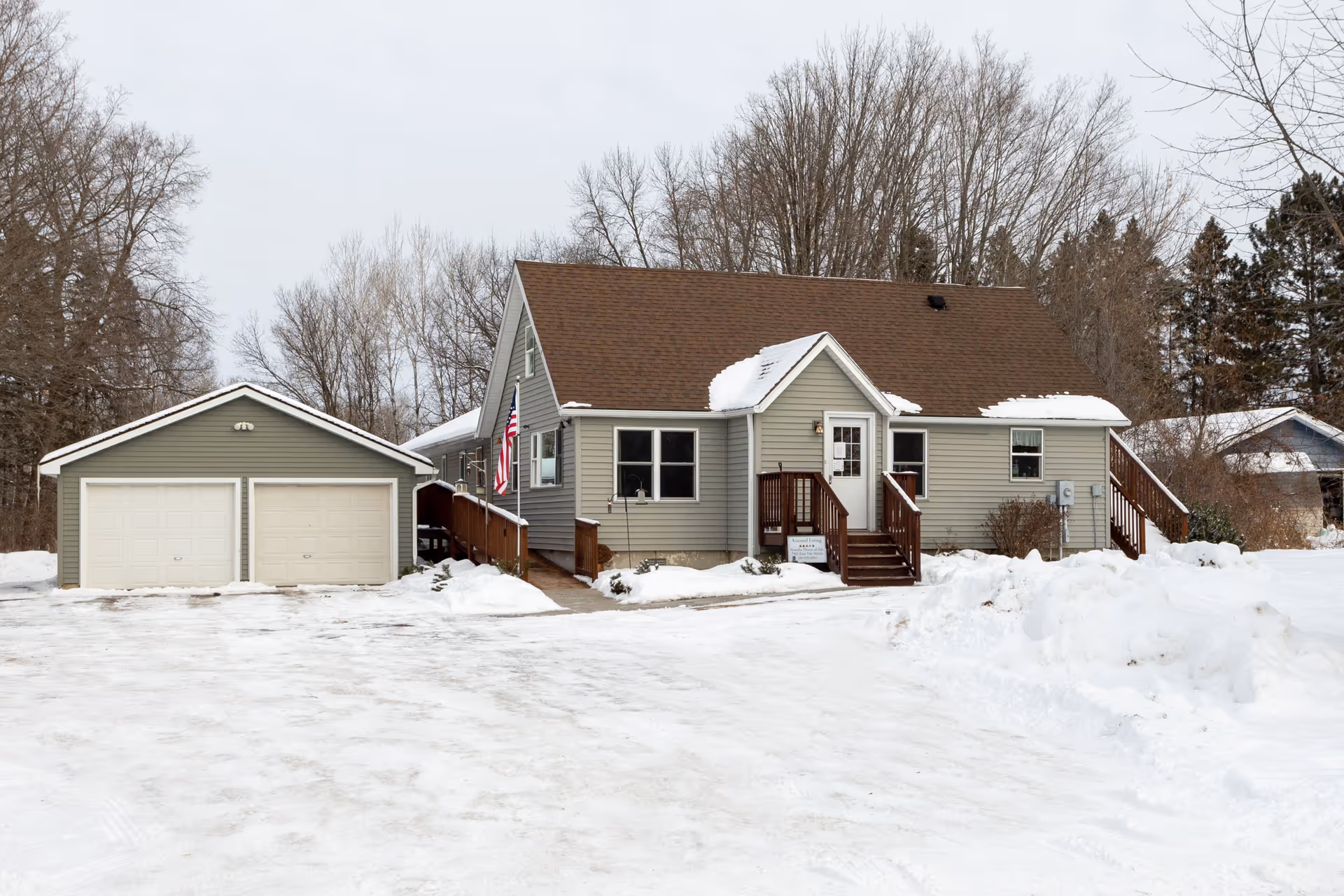 A single-story house with a brown roof and light gray siding surrounded by snow. There is a two-car garage attached on the left side, a wooden ramp and stairs leading to the front door, and leafless trees in the background under an overcast sky.