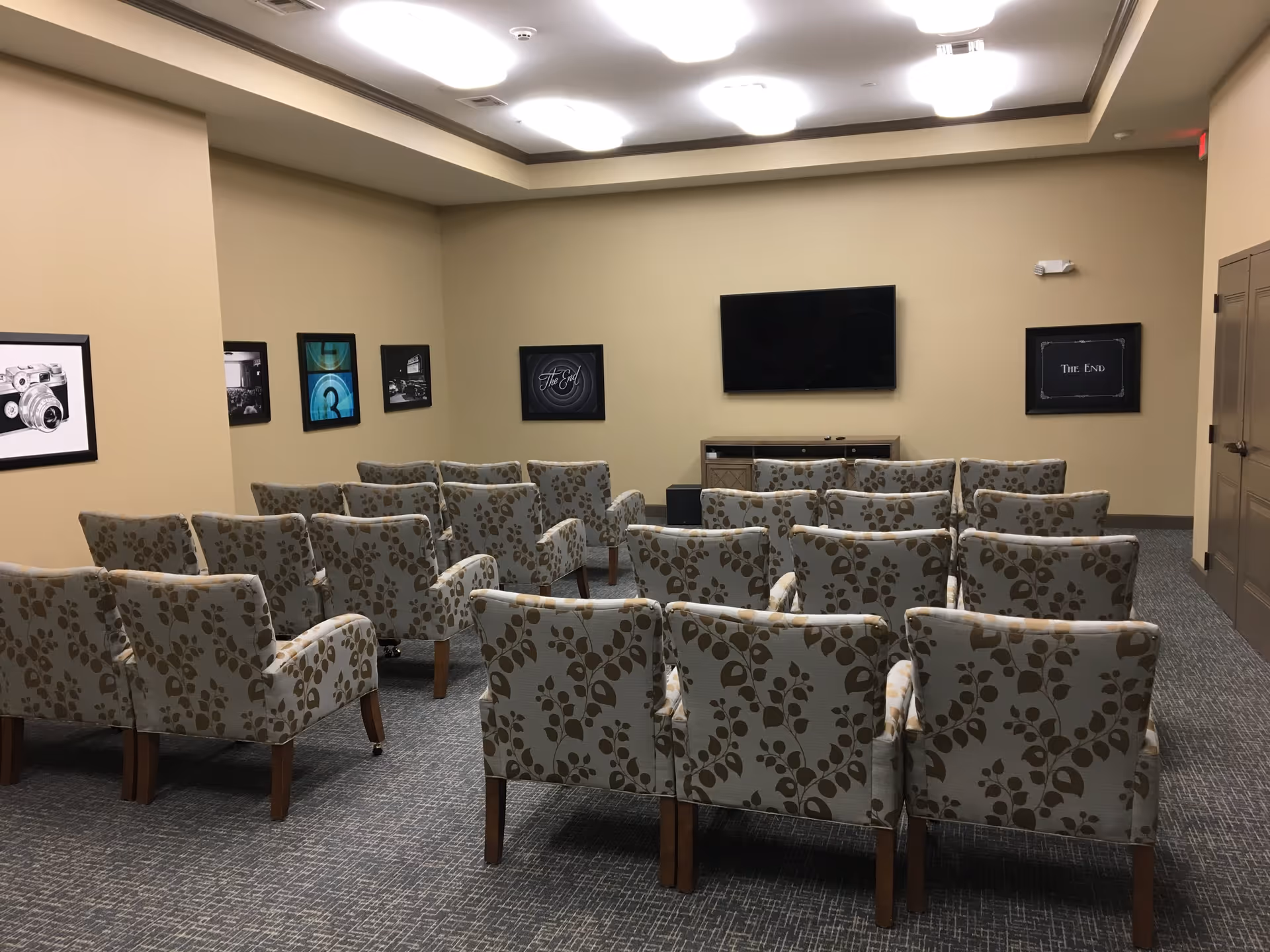 A small theater-style room with several rows of patterned armchairs facing a flat-screen TV mounted on a beige wall. The room has framed black and white pictures on the walls, a carpeted floor, and ceiling lights.