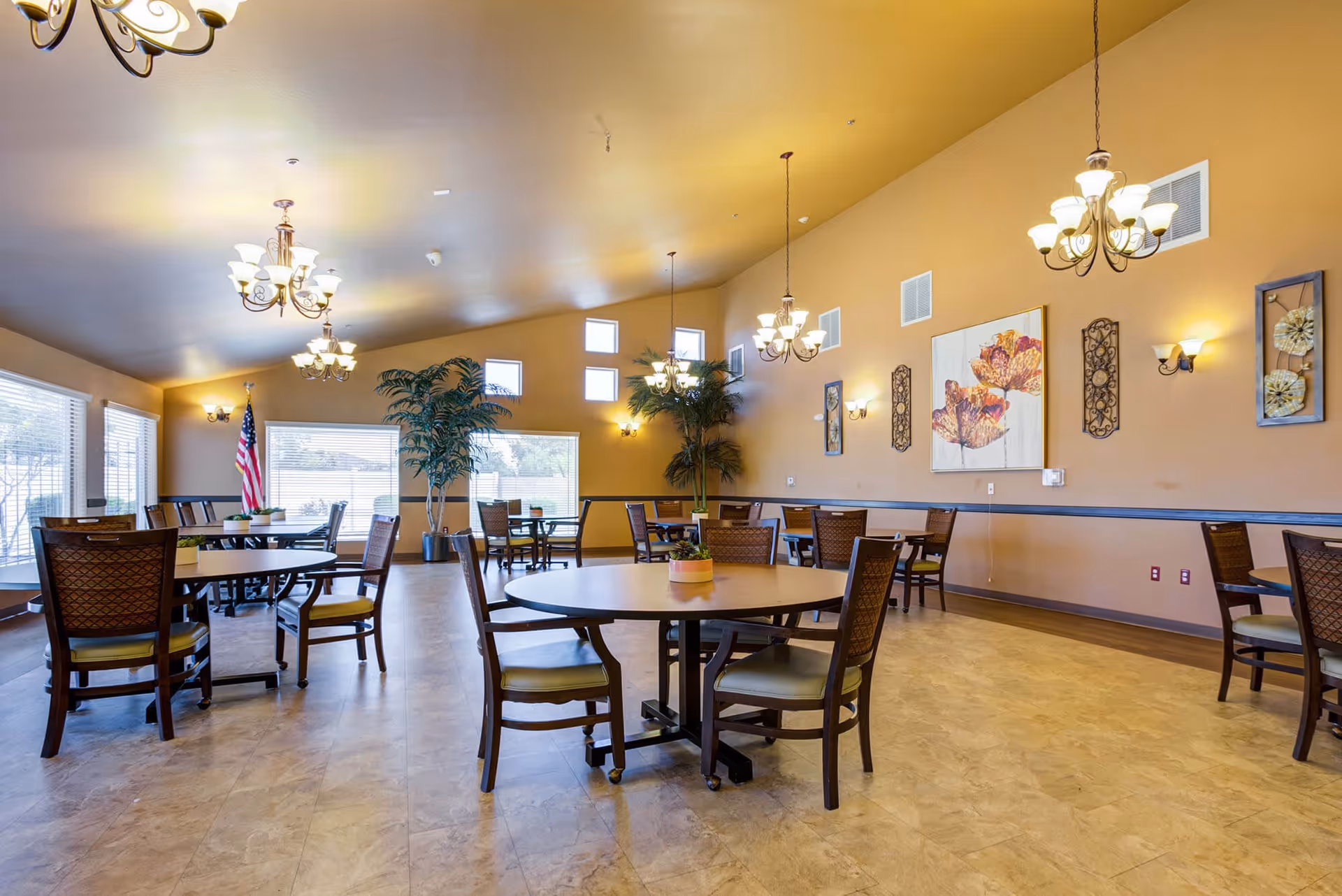 A spacious dining room with multiple round tables and wooden chairs with cushioned seats. The room has beige walls adorned with floral artwork and decorative wall sconces. Several chandeliers hang from the ceiling providing warm lighting. Large windows allow natural light to enter, and there are tall potted plants near the windows. An American flag is positioned near one of the windows.