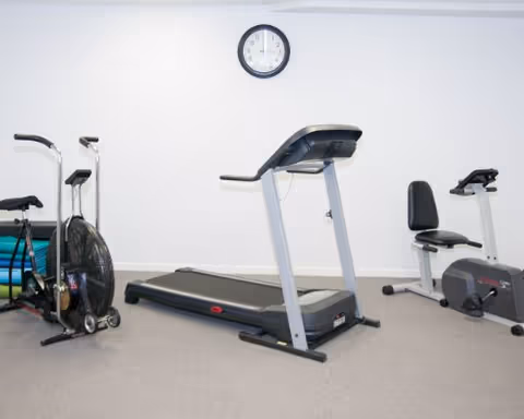 A small exercise room with a treadmill, a stationary bike, and an air bike against a plain white wall with a clock showing 11:10.