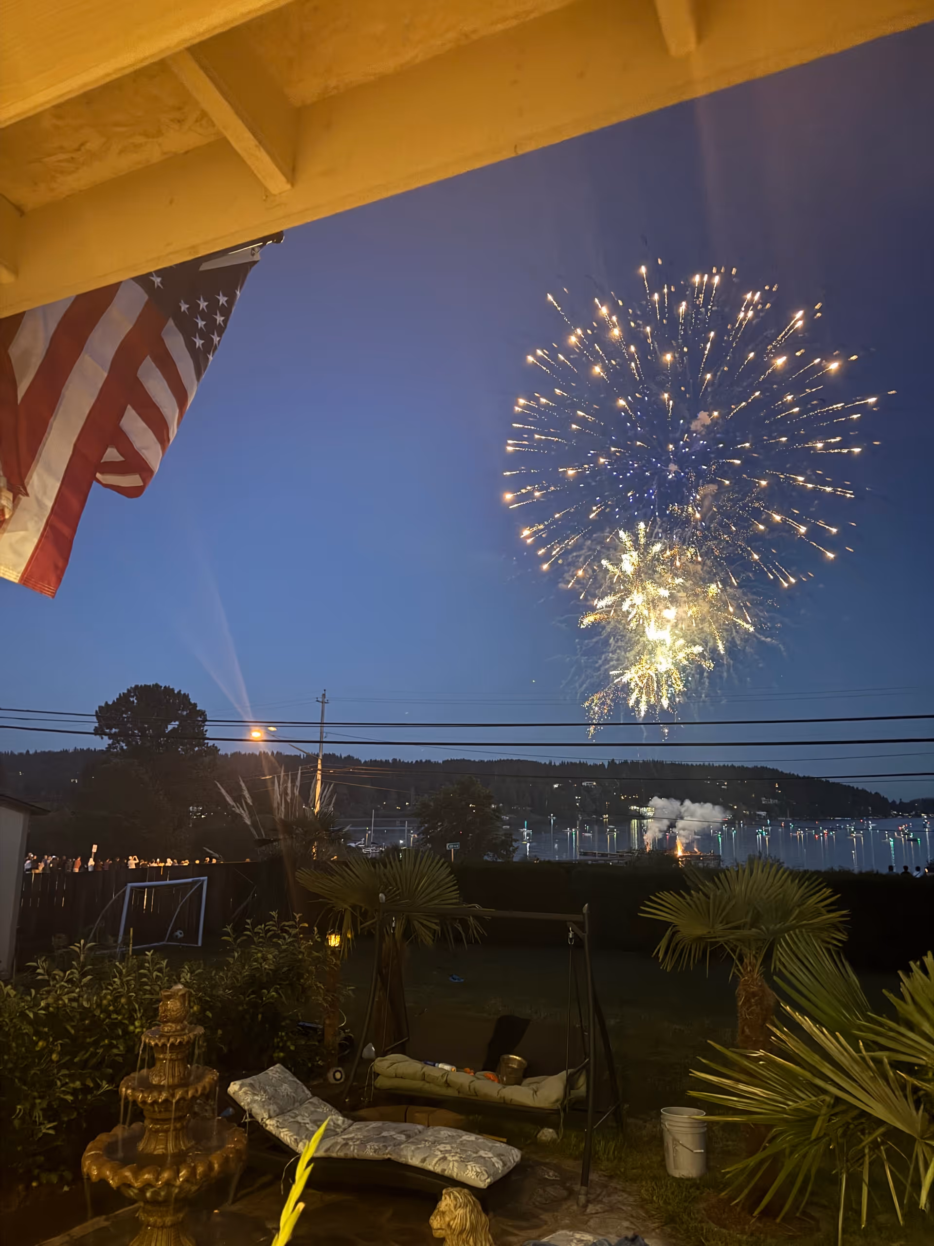 Fireworks burst over a waterfront at dusk, seen from a backyard patio with an American flag, lounge chairs, a fountain, and palm plants.