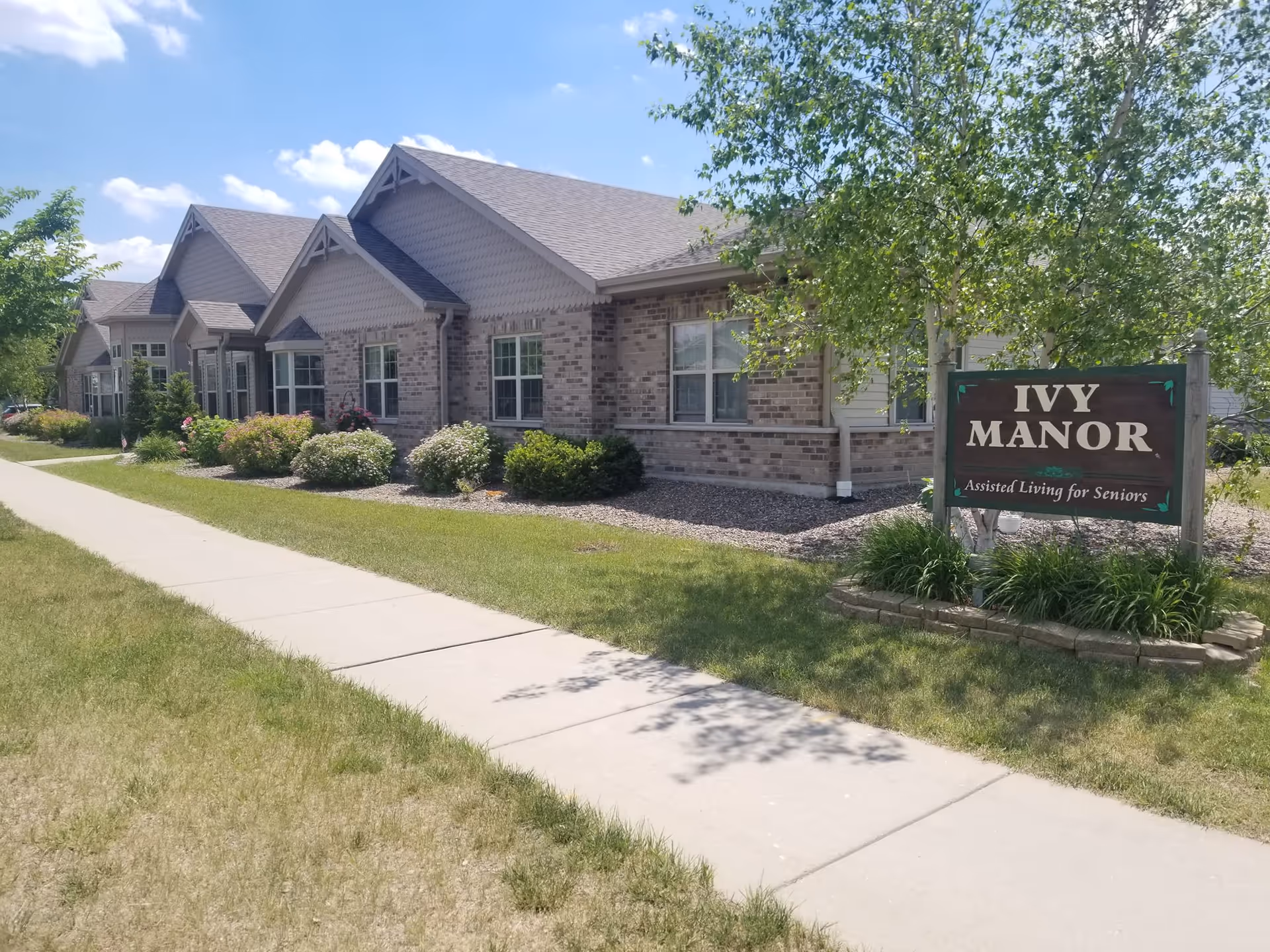 Exterior view of a single-story brick building with multiple gabled roofs, surrounded by green grass, bushes, and trees under a blue sky with some clouds. A sidewalk runs alongside the building. A wooden sign in front reads 'IVY MANOR Assisted Living for Seniors.'