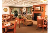 A cozy dining room in a senior living facility featuring round wooden tables set with plates and cups, comfortable upholstered chairs, a fireplace with a framed painting above it, large windows with curtains, a clock on the wall, and decorative plants.
