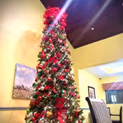 Tall Christmas tree decorated with red poinsettias, gold ornaments, and lights in a room with yellow walls, a wooden ceiling, and a dining area with chairs and framed artwork on the walls.