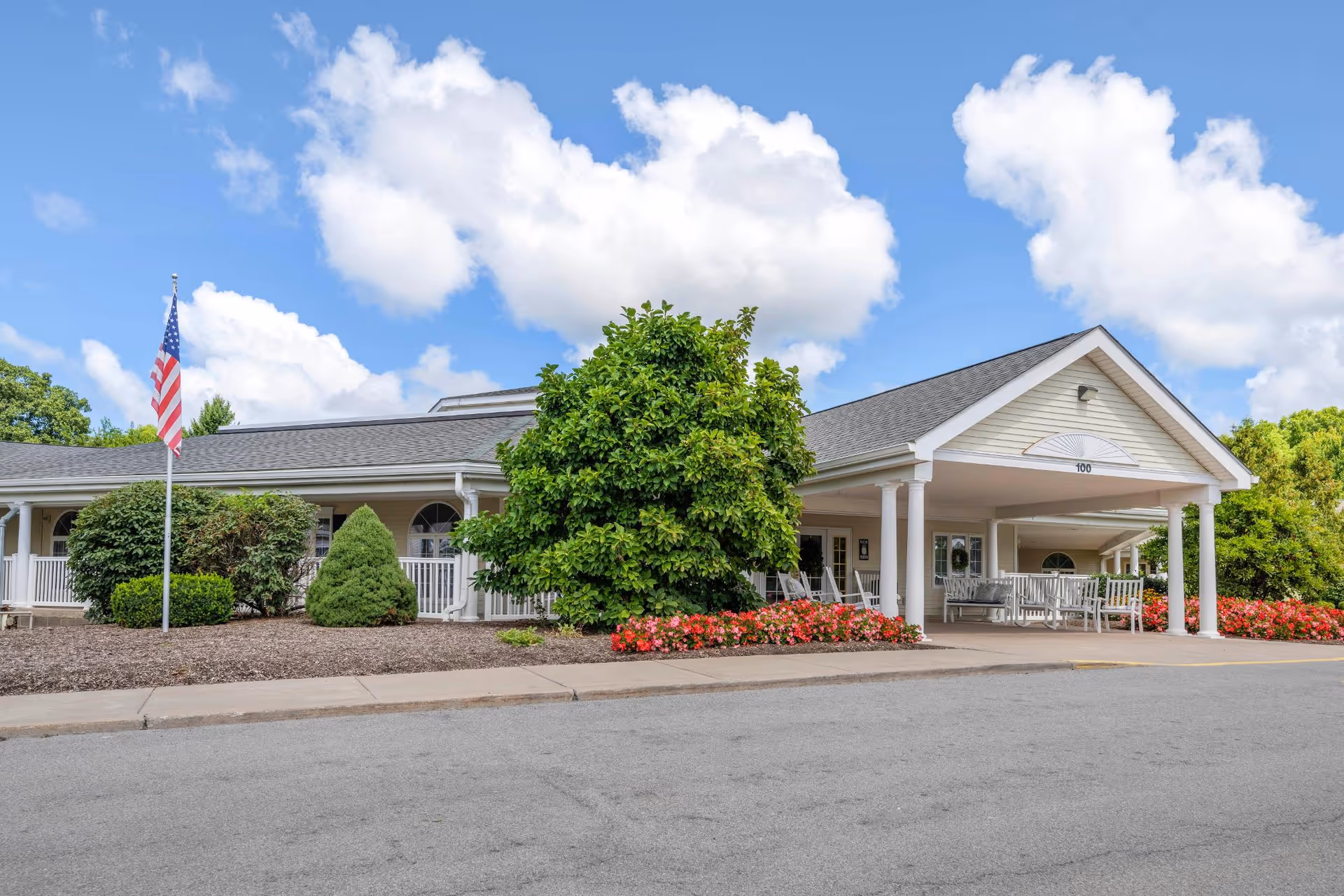 Front entrance of a single-story senior living facility with a covered drop-off, rocking chairs, landscaped shrubs and an American flag.