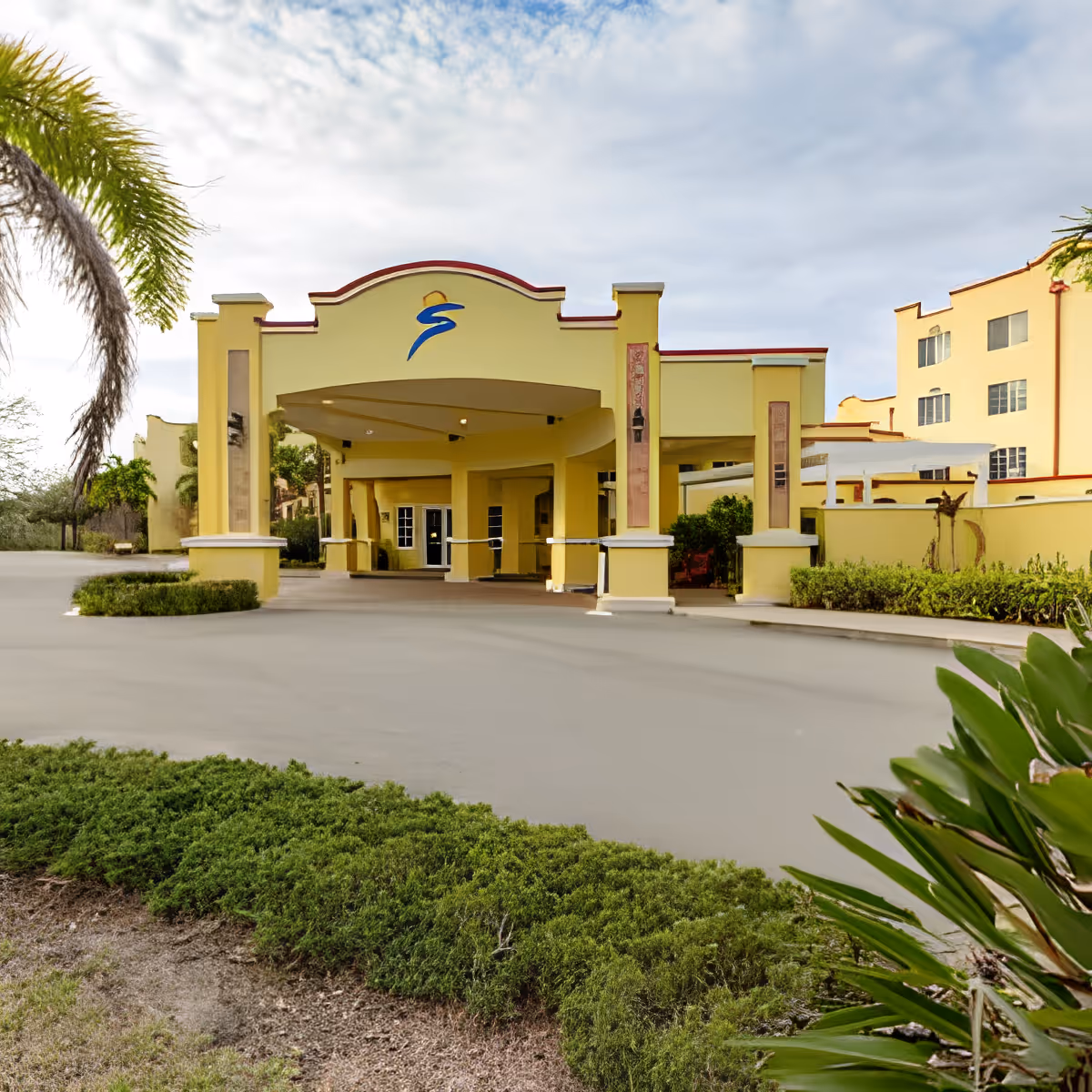 Exterior view of a yellow building with a covered entrance and columns, surrounded by greenery and palm trees under a partly cloudy sky.