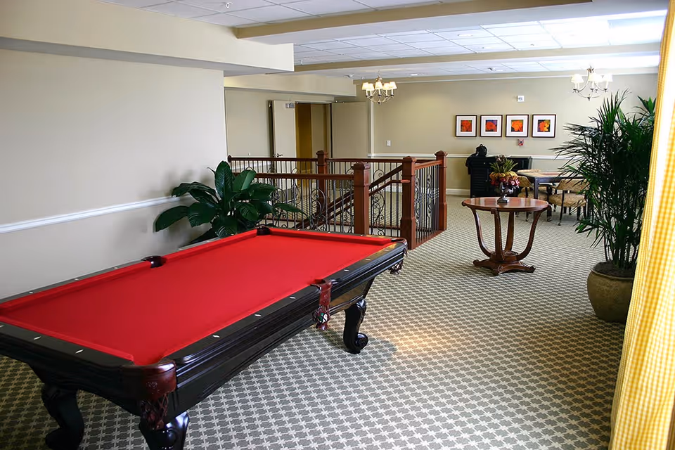 Common area in a senior living facility featuring a red-felt pool table, seating, plants, and decorative railing.