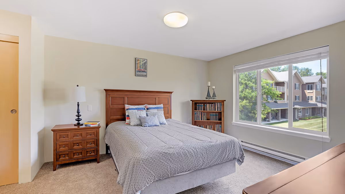A bright bedroom with a large window showing a view of neighboring buildings and greenery. The room features a wooden bed with a gray patterned comforter and multiple pillows, a wooden nightstand with a lamp and books, a wooden bookshelf filled with books, and a framed picture on the wall above the bed.