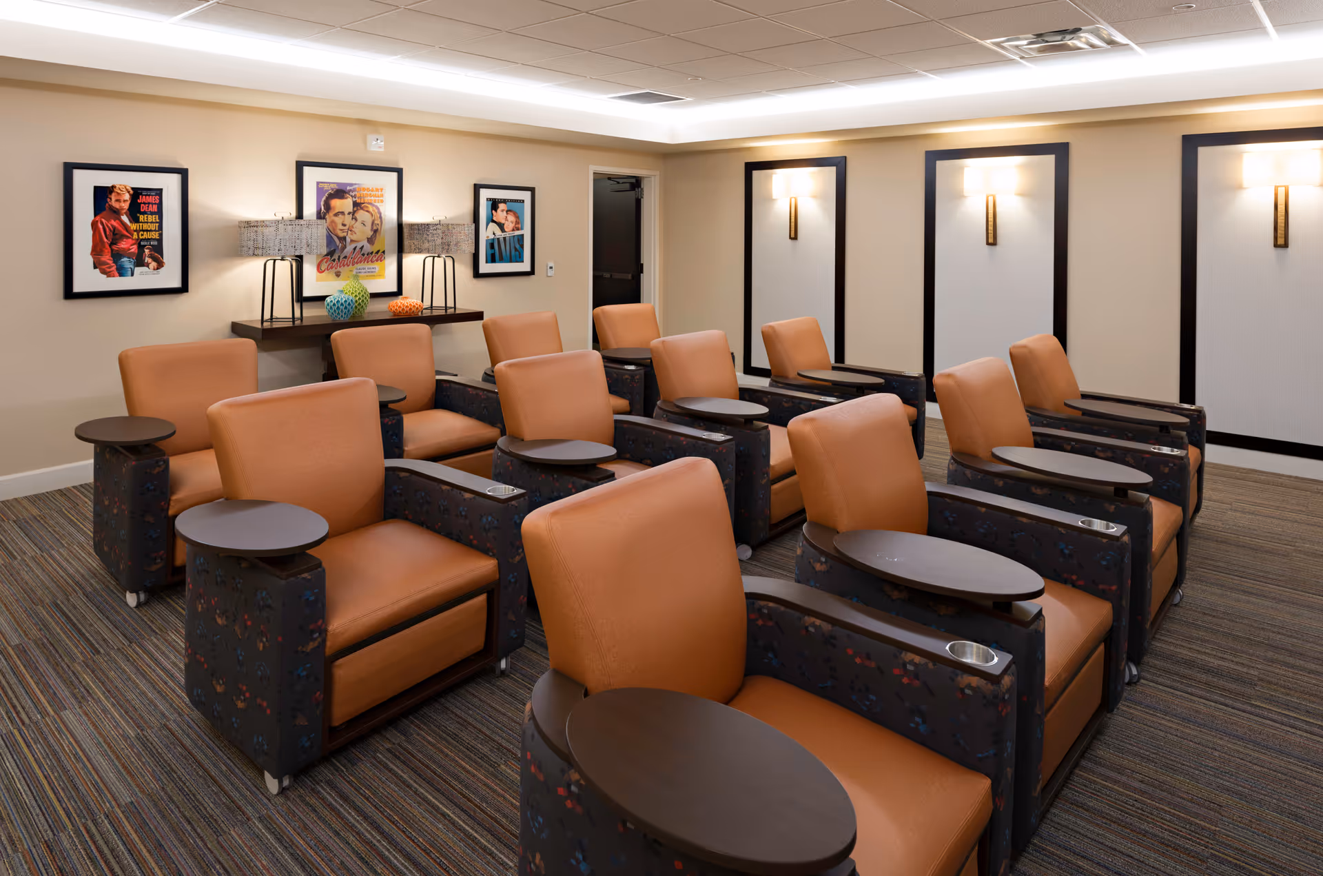 A small theater room with two rows of comfortable brown leather chairs, each equipped with a small attached table and cup holder. The walls are decorated with framed classic movie posters and modern wall sconces providing soft lighting. The room has a carpeted floor with striped patterns and a ceiling with recessed lighting.