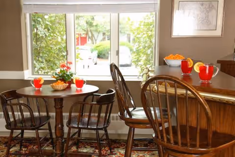 A cozy dining area with a round wooden table and four wooden chairs. On the table are three glasses of red beverage garnished with lemon slices and a bowl of snacks. Next to the table is a wooden counter with two high wooden chairs, a bowl of orange snacks, and two more glasses of the same red beverage. A large window behind the table shows greenery and a parked car outside.