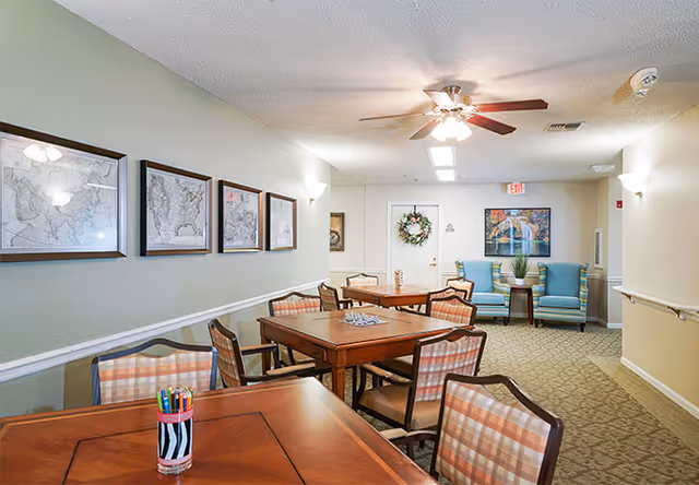 A well-lit common area in a senior living facility with several wooden tables and cushioned chairs arranged for group activities. The walls are decorated with framed maps and a large colorful painting. Two blue armchairs with a small table and plant are positioned near a door adorned with a wreath. The ceiling has a fan with lights, and the floor is carpeted.