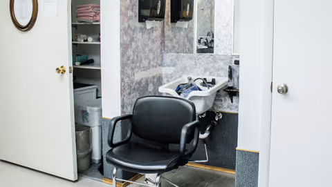 Interior view of a small hair salon or grooming area with a black salon chair in front of a white sink and mirror. To the left, there is an open door revealing shelves with folded towels and various supplies.