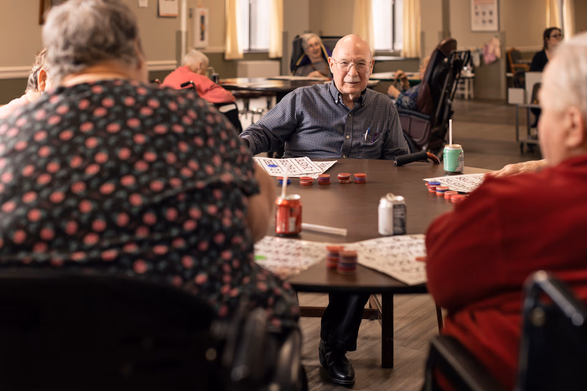 A group of elderly people sitting around a table playing bingo in a well-lit common area of a senior living facility. The man in the center is smiling and looking towards the camera, with bingo cards and chips on the table. Other residents are visible in the background, some in wheelchairs.
