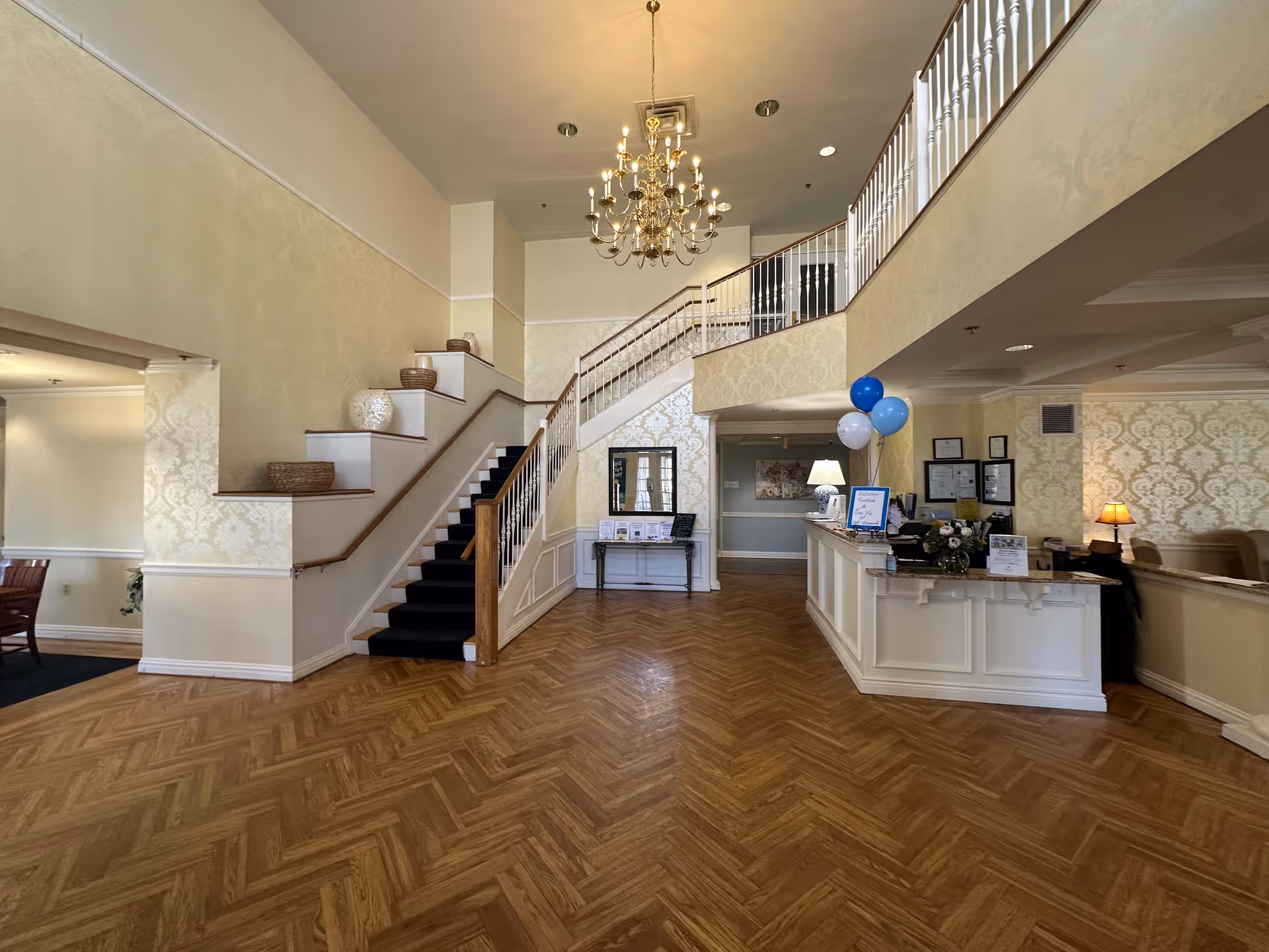 Spacious and elegant interior lobby area of Mira Vie at Clifton Assisted Living featuring a wooden herringbone floor, a grand staircase with a dark carpet runner, a chandelier hanging from the ceiling, a reception desk decorated with blue and white balloons, and patterned wallpaper on the walls.