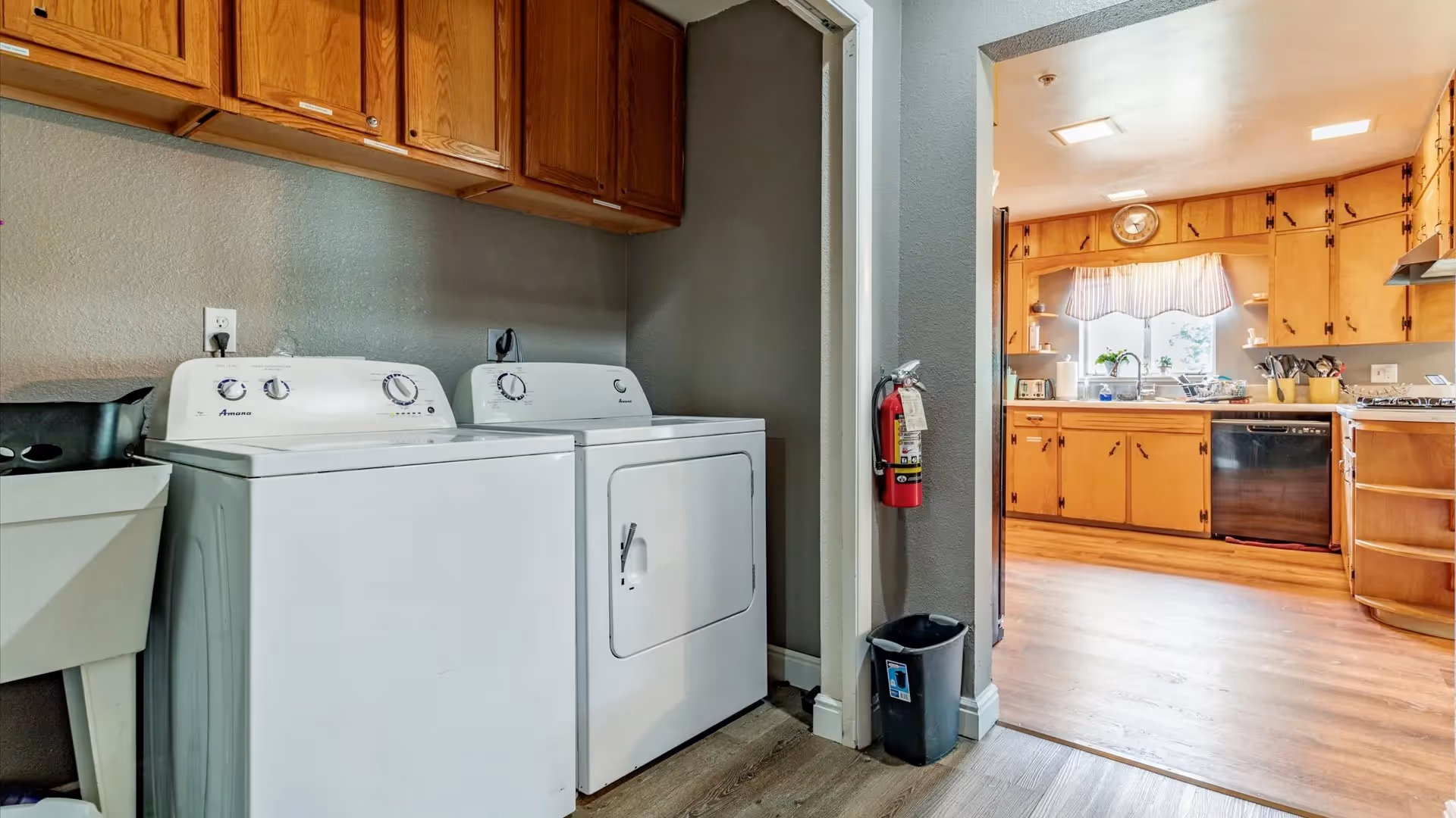 Laundry area with a white washing machine and dryer side by side, wooden cabinets above them, a utility sink to the left, and a fire extinguisher mounted on the wall next to an open doorway leading to a kitchen with wooden cabinets, a window with a valance, and various kitchen appliances.
