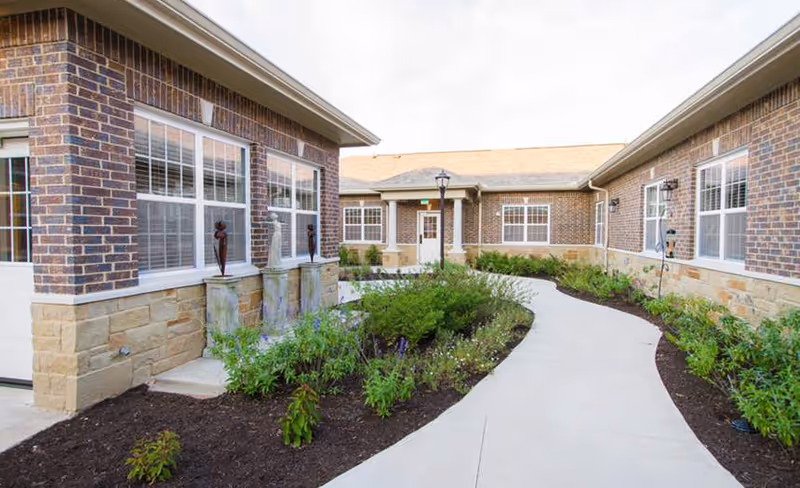 Curved concrete walkway through a landscaped courtyard between brick single-story memory care building wings.