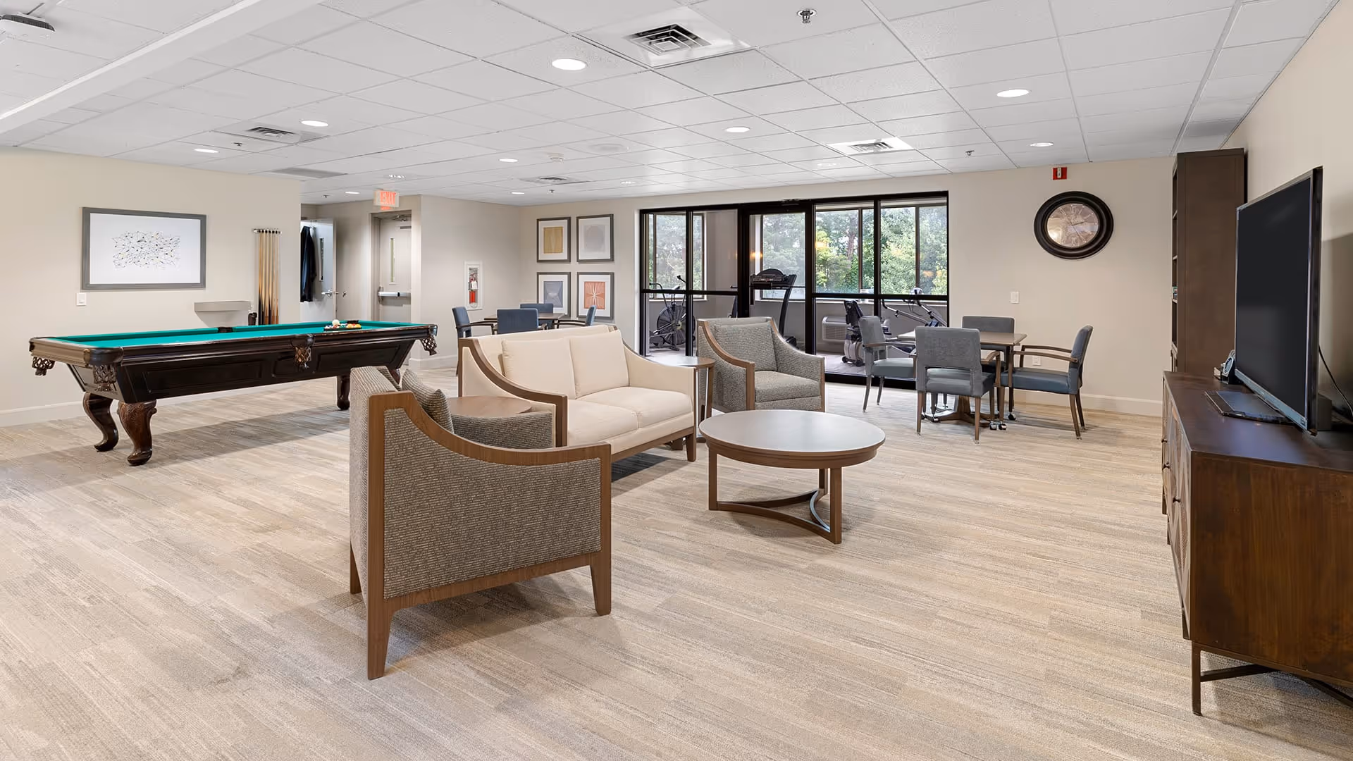 A spacious common area in a senior living facility featuring a pool table, a seating area with a sofa and two armchairs around a round coffee table, a television on a wooden stand, and several tables with chairs. Large windows and glass doors reveal an exercise room with fitness equipment outside the main room.