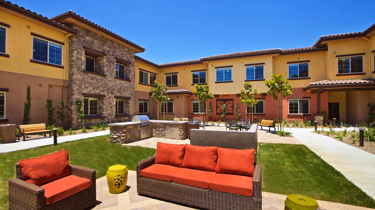 Outdoor courtyard area of a senior living facility with wicker patio furniture featuring red cushions, a built-in stone grill, green grass, small trees, and a two-story building with yellow and brown exterior walls under a clear blue sky.
