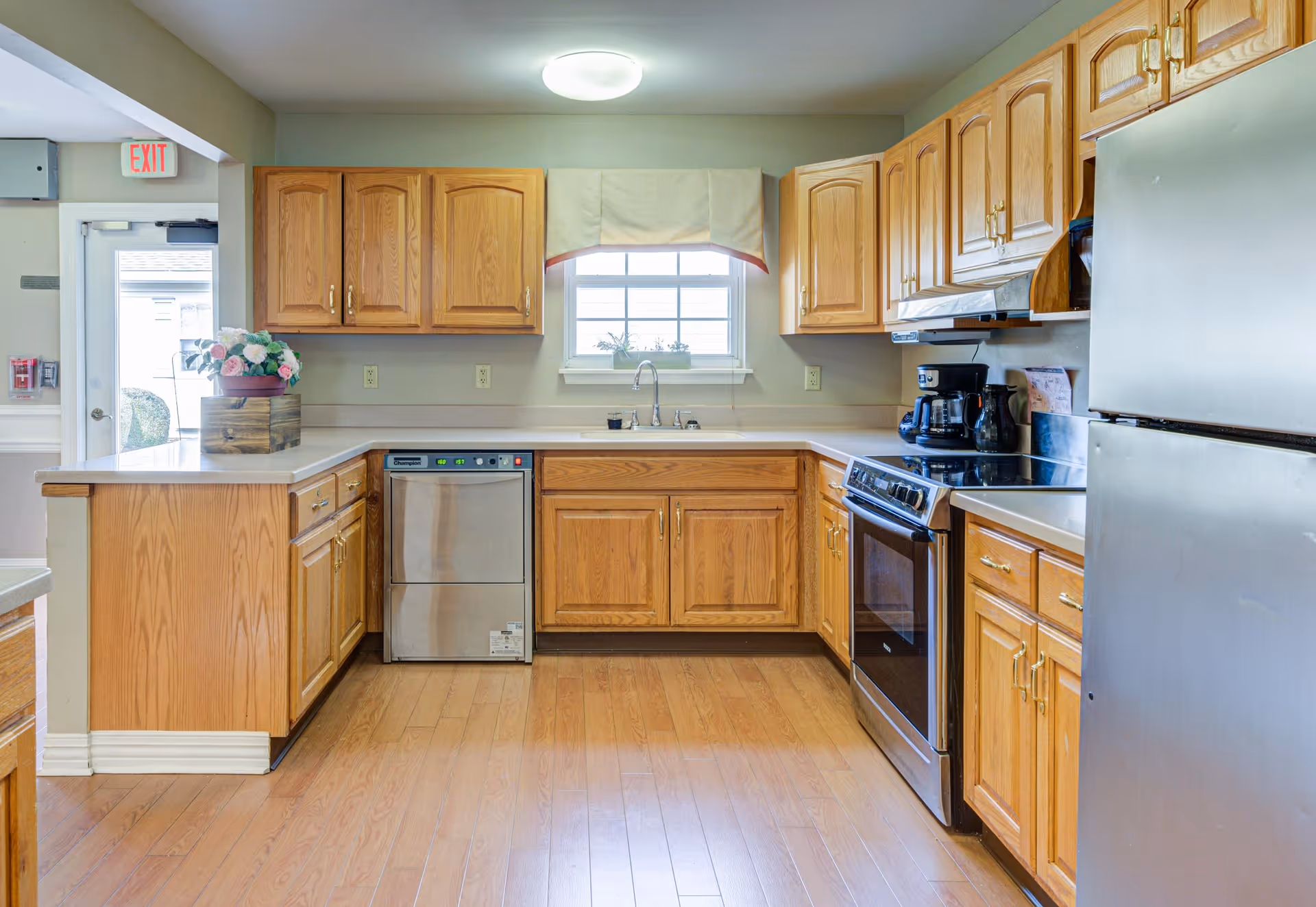 A bright kitchen with wooden cabinets, a stainless steel dishwasher, stove, and refrigerator. There is a window above the sink with a valance, a flower arrangement on the counter, and a door with an exit sign visible in the background.