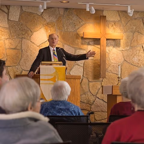 A man in a suit and colorful striped tie stands behind a wooden podium with a microphone, speaking to an audience of elderly people seated in chairs. Behind him is a stone wall with a large wooden cross mounted on it, and a lit candle on a small table to the side.