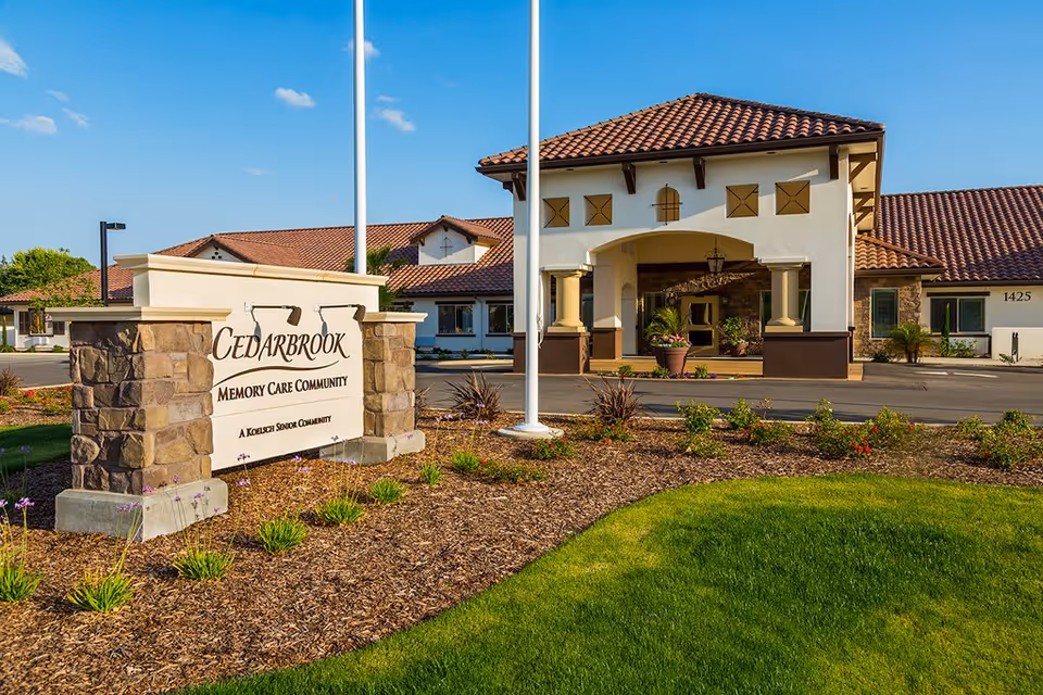 Exterior view of Cedarbrook Memory Care Community building with a stone and stucco sign in the foreground. The building has a tiled roof and an arched entrance with columns. There are landscaped plants and green grass around the sign and building under a clear blue sky.