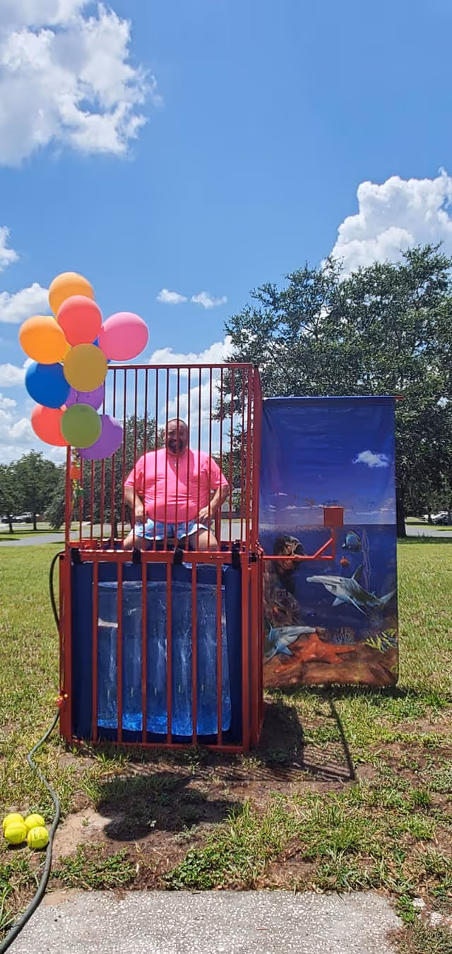 A man wearing a bright pink shirt sits inside a dunk tank cage outdoors on a sunny day. The dunk tank is decorated with colorful balloons on the left side and an underwater ocean scene with sharks on the right side. The area around the dunk tank is grassy with trees and a blue sky with clouds in the background.