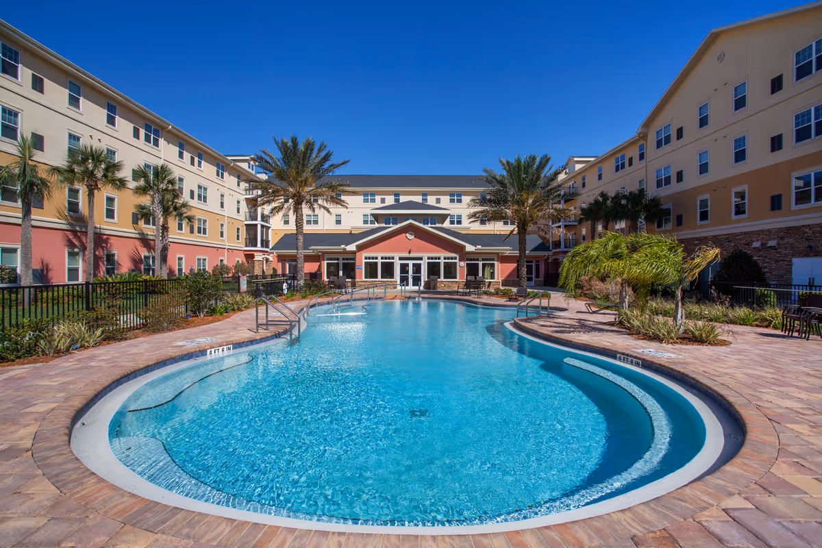 Outdoor swimming pool surrounded by a paved deck with lounge chairs and palm trees, set in the courtyard of a multi-story senior living facility building under a clear blue sky.