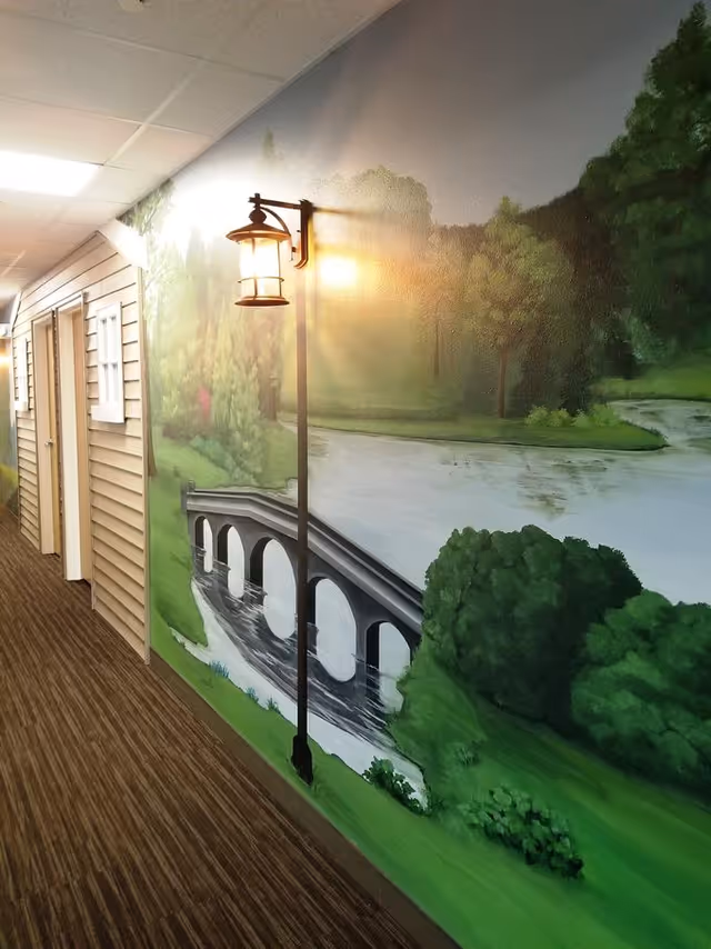 Hallway inside a senior living facility with a painted riverside mural showing a bridge and lamppost and doors along the corridor.