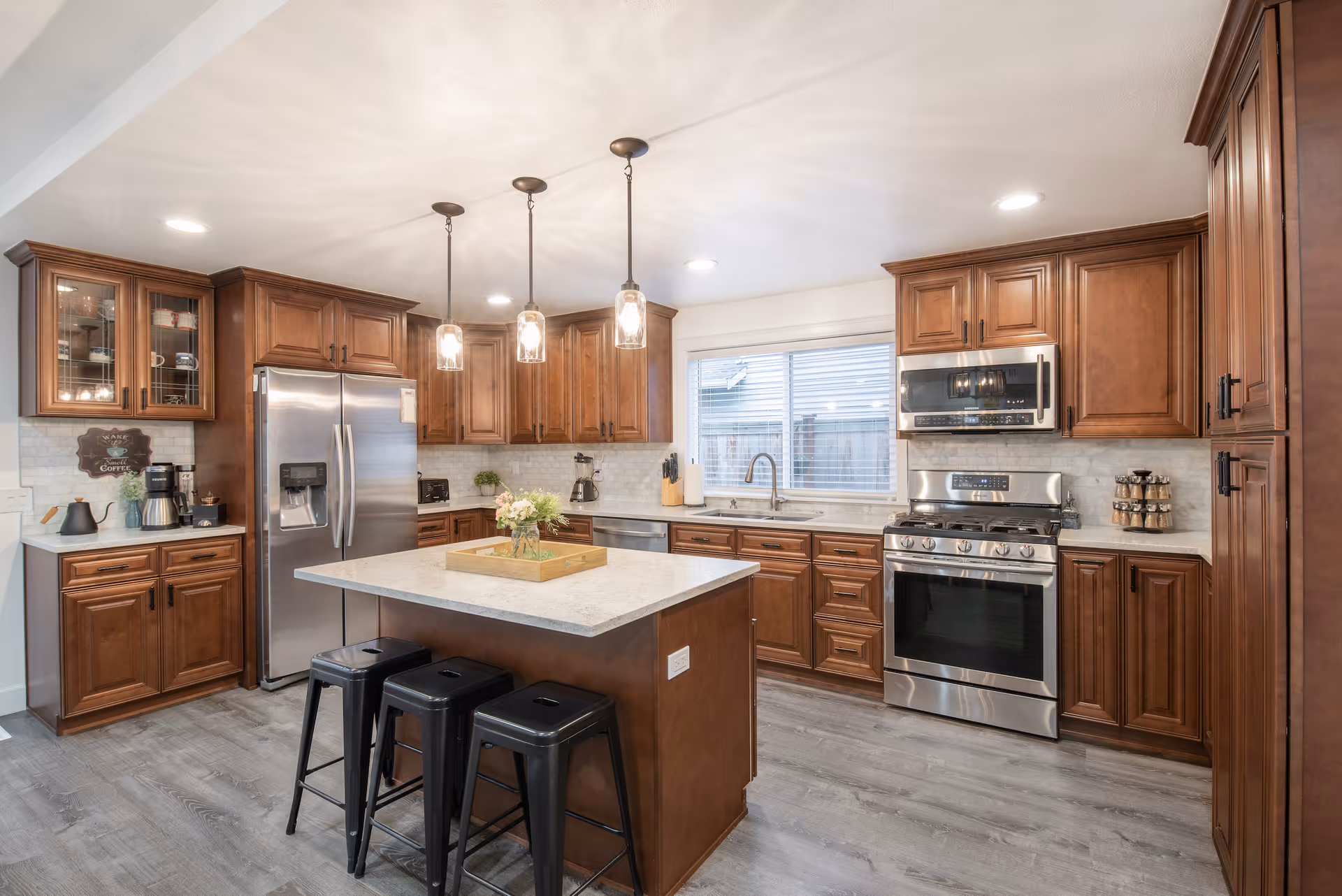 A spacious kitchen with wooden cabinets, a large island with a light countertop, three black stools, stainless steel refrigerator, stove, and microwave. The kitchen has pendant lights hanging above the island and a window above the sink letting in natural light.