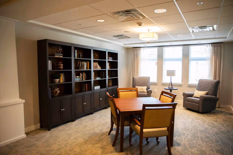 A cozy senior living common area with a wooden table and four chairs in the center, two comfortable armchairs with cushions near windows with curtains, a side table with a lamp between the armchairs, and a large dark wooden bookshelf filled with books and decorative items against the wall.