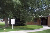 Brick building entrance with a sidewalk, lawn, large tree, and a sign on the grass.