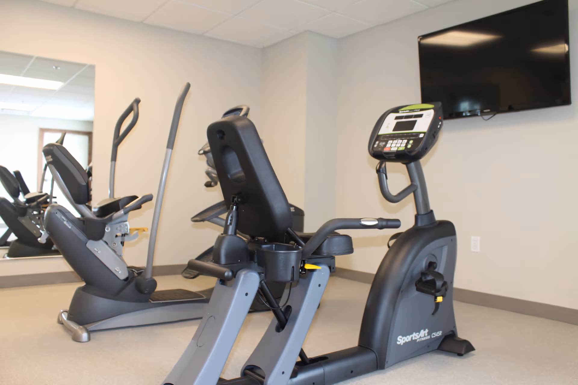 Indoor fitness room with exercise equipment including a recumbent stationary bike and an elliptical machine. A large mirror covers one wall, and a flat-screen TV is mounted on another wall.
