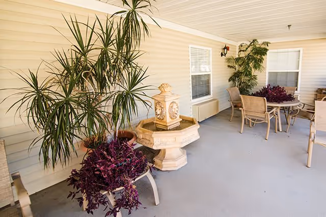 Covered outdoor patio area with beige siding walls, featuring a stone water fountain, several potted plants, and a round table with four chairs. The space is well-lit with natural light coming through two windows.