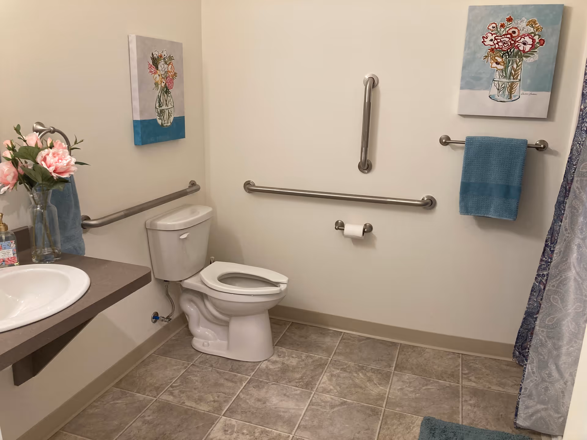 A clean bathroom with a white toilet, beige tiled floor, and white walls. There are stainless steel grab bars on the walls around the toilet for accessibility. A white sink with a brown countertop is visible on the left side with a vase of pink flowers and a soap dispenser. Two floral paintings hang on the walls, and blue towels are placed on towel racks. A patterned shower curtain is partially visible on the right side.