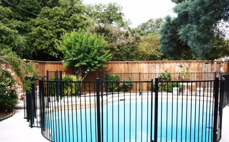 Black metal-fenced swimming pool in a landscaped backyard with trees and a wooden privacy fence.