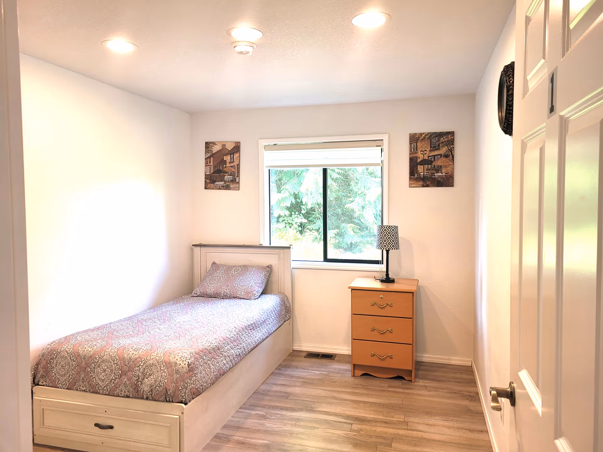 A small, bright bedroom with a single bed covered in a patterned bedspread and a matching pillow. Next to the bed is a wooden nightstand with three drawers and a decorative lamp on top. The room has light-colored walls, wood flooring, a window with a view of greenery outside, and two framed pictures hanging on the wall beside the window. The door to the room is partially open.
