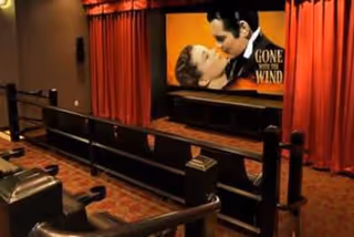 Interior of a small movie theater room with rows of black seats and a large screen displaying the movie poster for 'Gone with the Wind'. The room has red curtains on the sides of the screen and warm lighting.