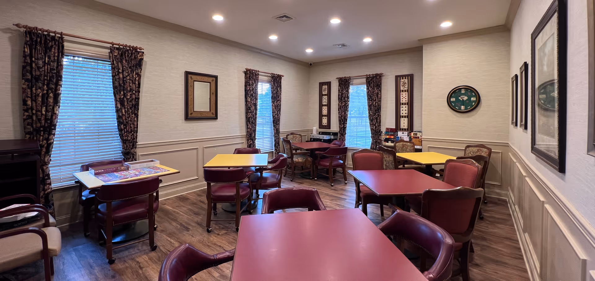 A cozy common room with several tables and chairs arranged for group activities or dining. The room has wood flooring, beige walls with wainscoting, and windows with floral curtains. There are board games and puzzles on some tables, framed artwork on the walls, and a clock shaped like a poker table.