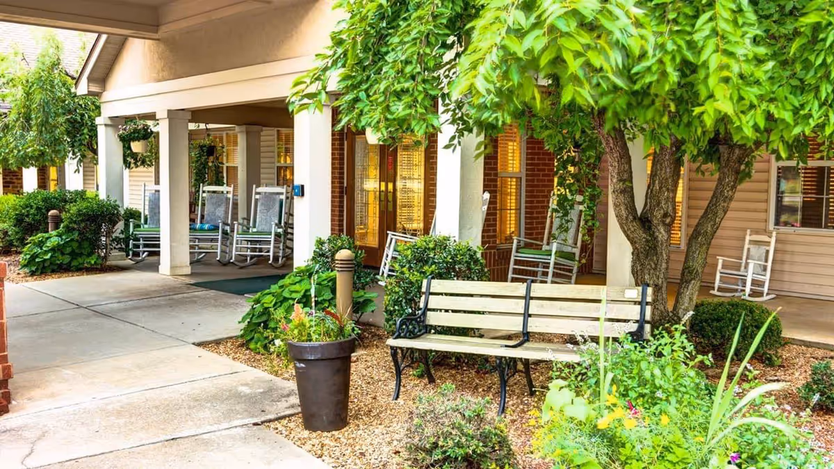 Covered front porch of an assisted living community with rocking chairs, a bench, potted plants, and a tree.