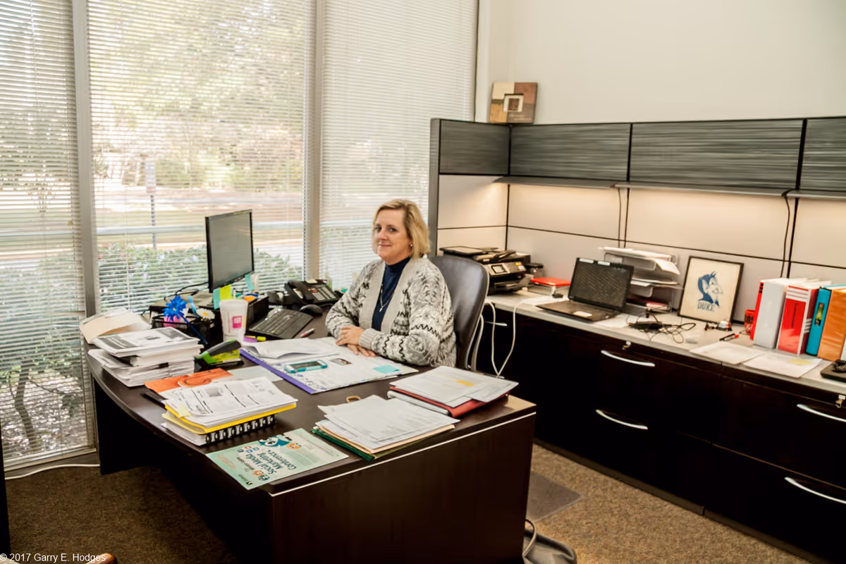A woman sits at a cluttered office desk with papers, computer monitors, and filing cabinets in a bright workspace with large windows.