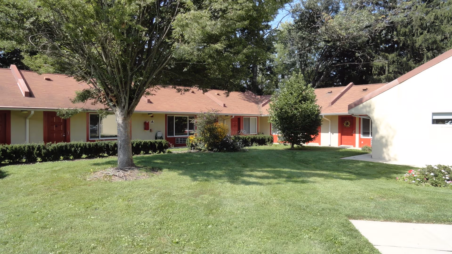 Single-story building with red doors and beige walls surrounding a grassy courtyard with trees and shrubs under a clear sky.