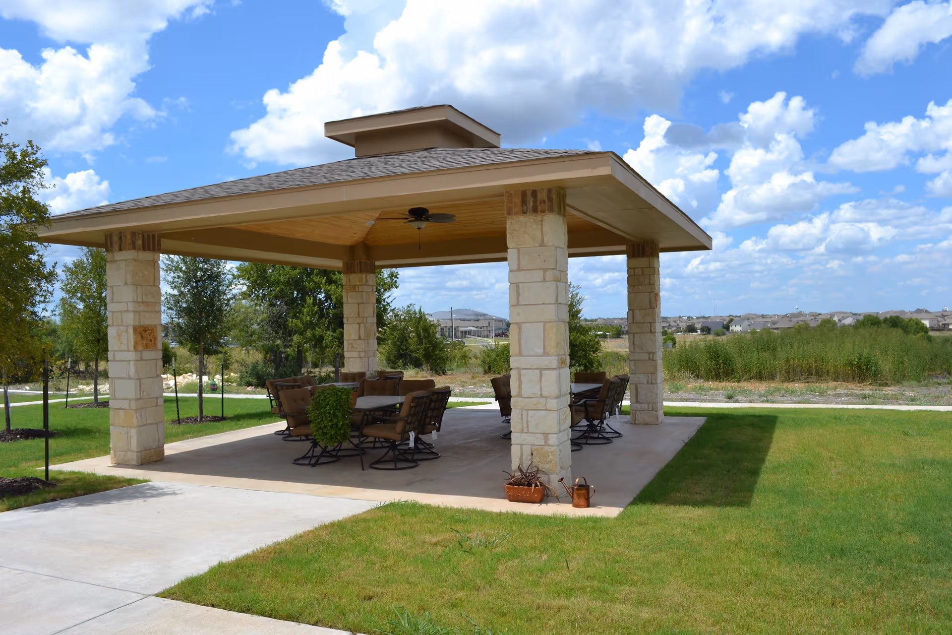 Outdoor covered pavilion with stone pillars and a wooden ceiling, furnished with multiple cushioned chairs and tables, surrounded by green grass and trees under a partly cloudy blue sky.