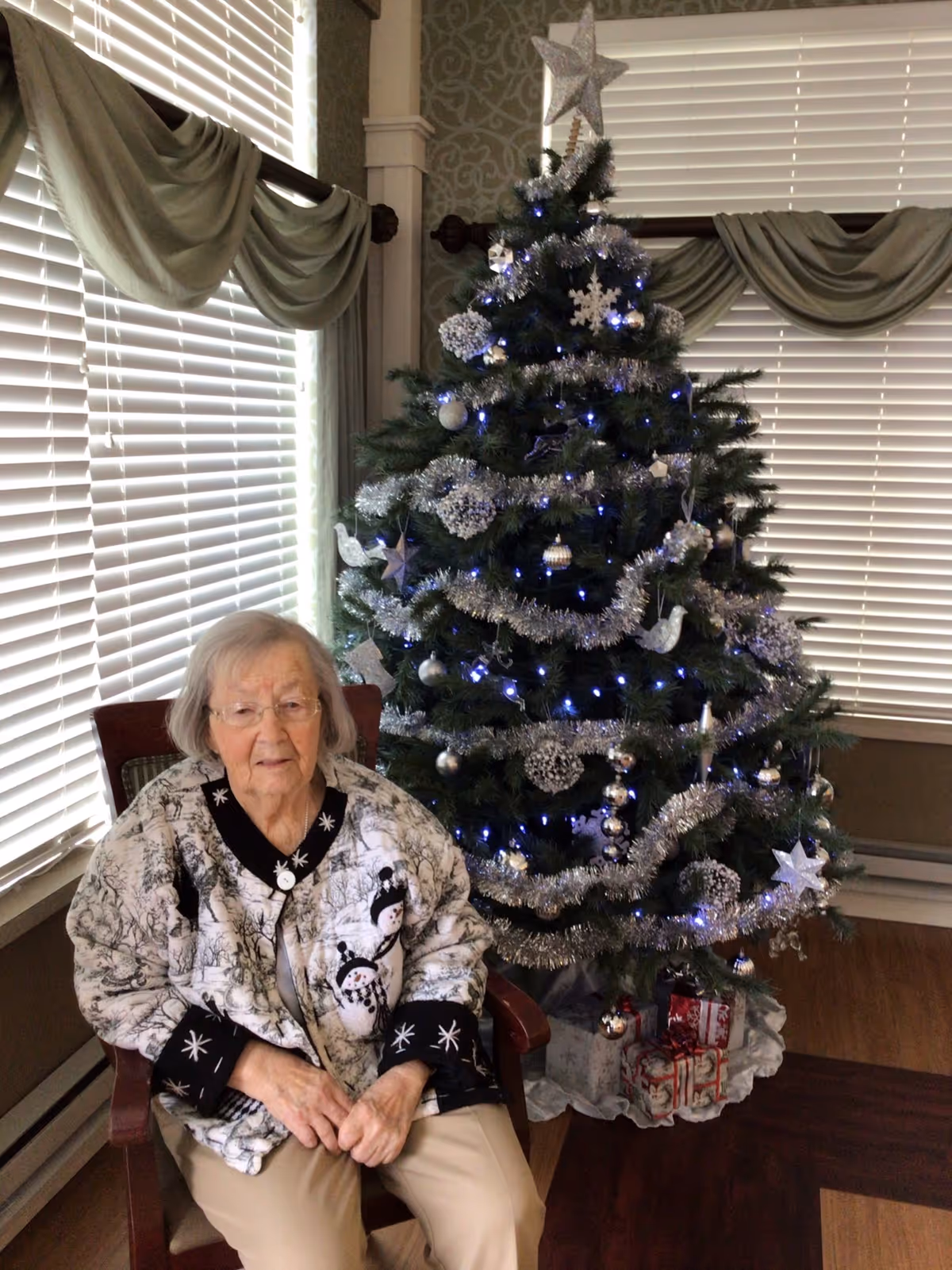 An elderly woman with gray hair and glasses sitting on a wooden chair next to a decorated Christmas tree with silver garlands, ornaments, and blue lights in a room with large windows covered by white blinds and green drapes.