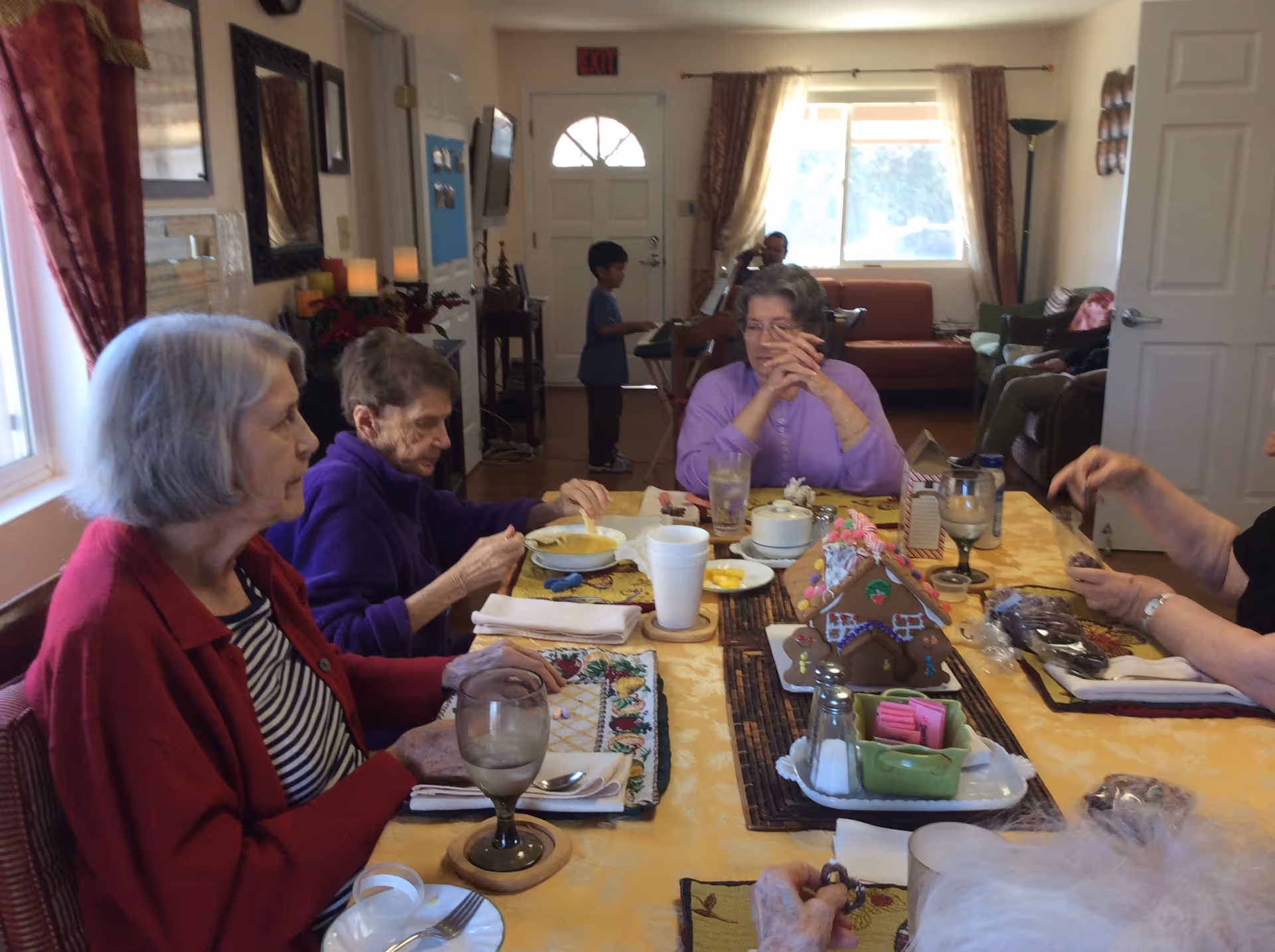 A group of elderly women sitting around a dining table in a residential care facility, engaging in conversation and eating. The table is set with placemats, utensils, glasses, and a decorative gingerbread house centerpiece. In the background, a young boy stands near the entrance door, and a few other people are seated in a living room area with couches and a window letting in natural light.