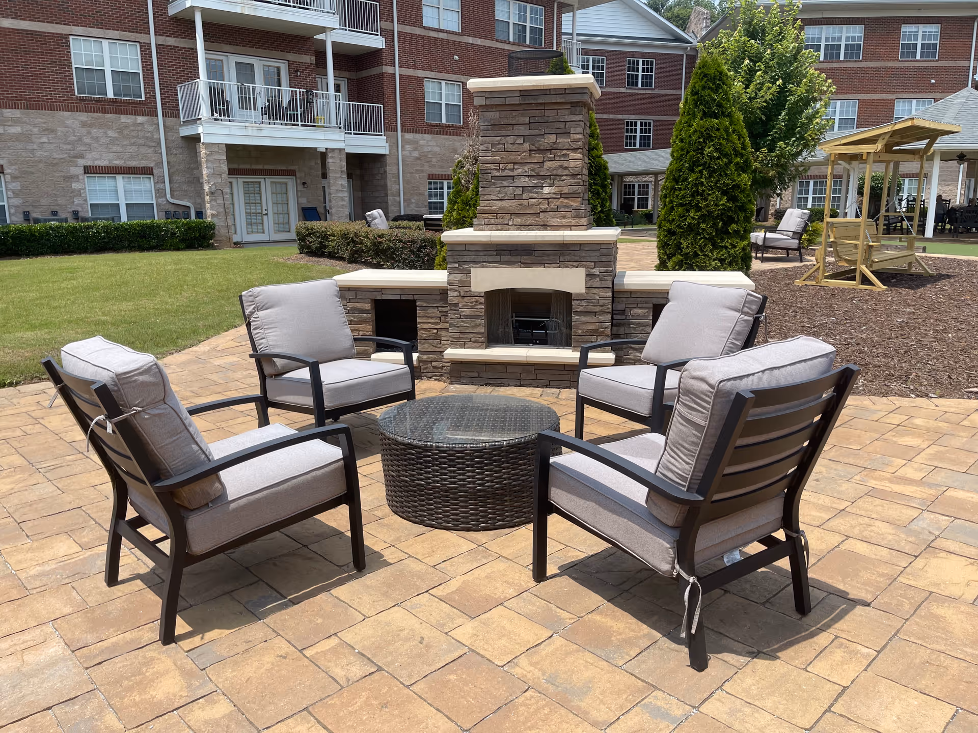 Outdoor patio area with four cushioned chairs arranged around a round wicker table in front of a stone fireplace. The setting is part of a senior living facility with brick buildings, green lawn, and a wooden swing in the background.