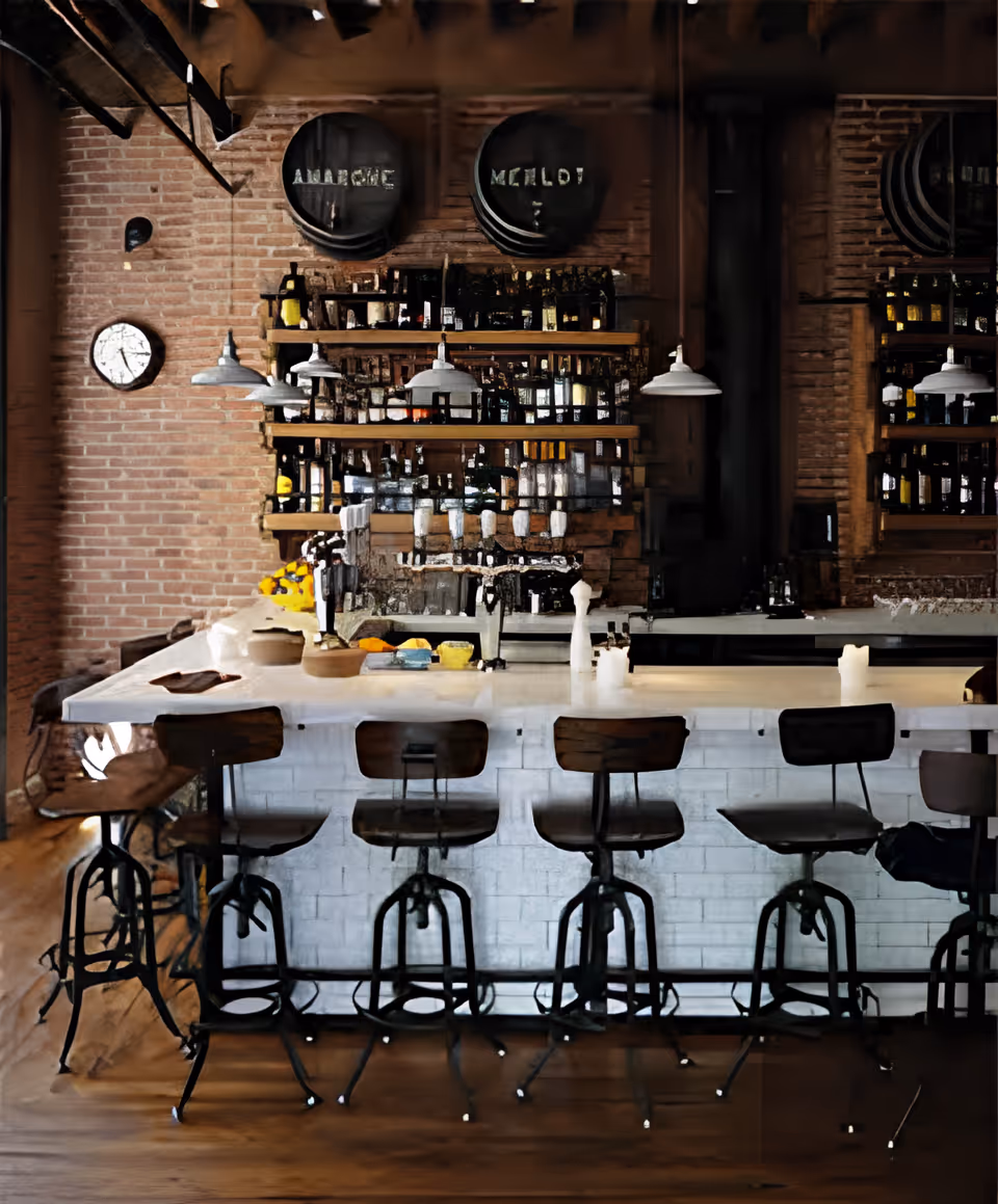 Interior view of a bar area with a white countertop and six wooden bar stools with metal legs. Behind the bar, there are shelves stocked with various bottles and glassware against a brick wall. Three industrial-style pendant lights hang above the bar, and a round clock is mounted on the brick wall to the left.