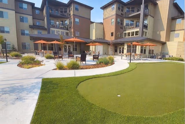Outdoor patio area of a senior living facility with tables, chairs, and orange umbrellas surrounded by landscaped plants and a putting green in the foreground. The multi-story building with balconies and large windows is visible in the background under a clear sky.