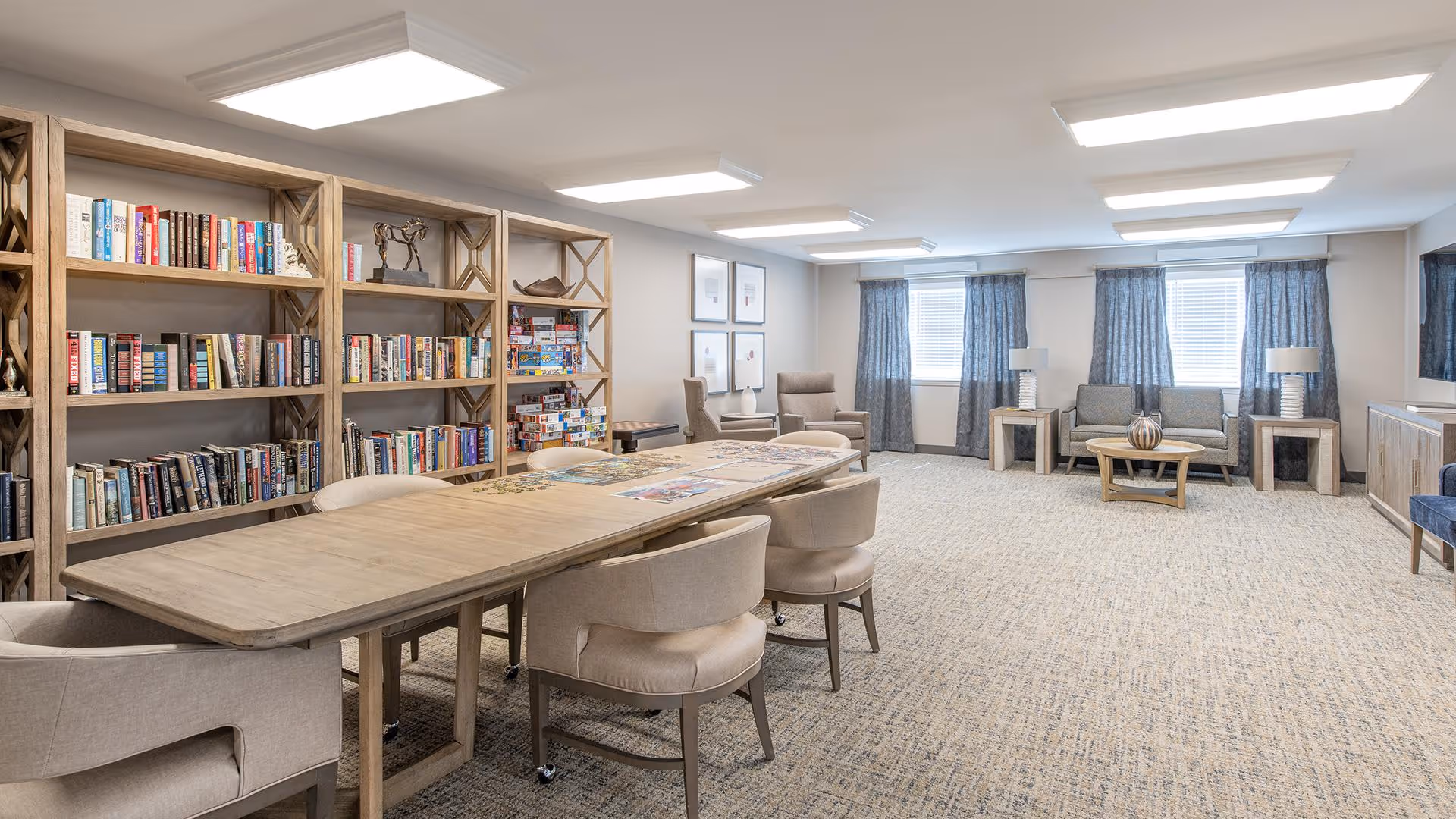A spacious, well-lit common room with a large wooden table surrounded by beige upholstered chairs. Along one wall, there are wooden bookshelves filled with books and board games. The room also features a sitting area with a sofa, armchairs, side tables with lamps, and windows with blue curtains. The carpet is light-colored and the ceiling has recessed lighting panels.