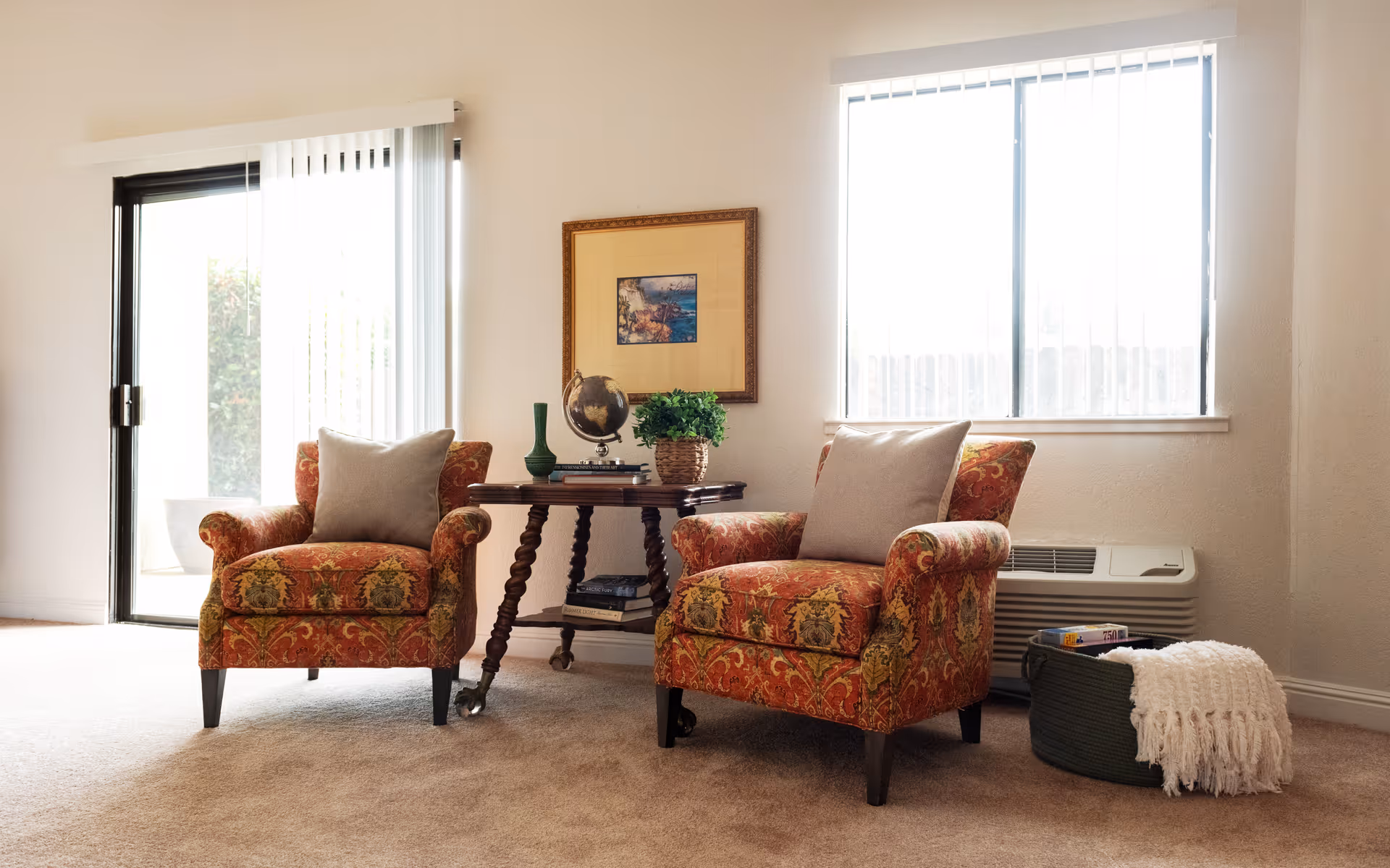Two patterned upholstered armchairs with throw pillows flank a small wooden side table in a bright living room with a sliding glass door and window.