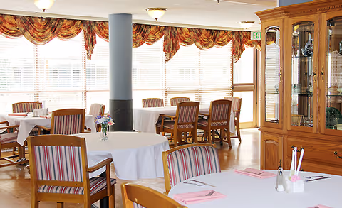 Bright dining room with round tables set with white tablecloths, striped wooden chairs, large windows with patterned valances, and a china cabinet.